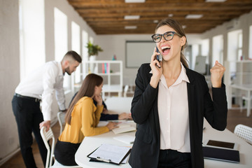 Young business woman in eyeglasses and shirt screaming while talking on cellphone in office with colleagues on background