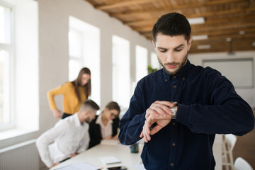 Young man with beard in shirt thoughtfully looking on handwatch in office with colleagues on background