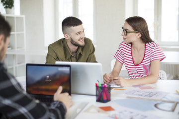 Young man in shirt and woman in striped t-shirt and eyeglasses happily discussing work moments together. Group of cool guys working with laptop while spending time in modern office