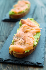 Rye toasts with smoked salmon and mashed avocado served on slate plates. Dark wooden background, high resolution