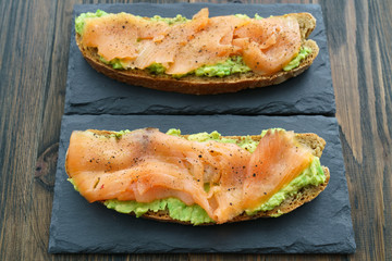 Rye toasts with smoked salmon and mashed avocado served on slate plates. Dark wooden background, high resolution