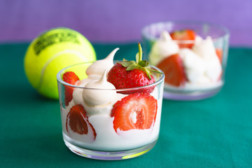 Wimbledon inspired whipped cream, meringues and fresh strawberries in a glass bowl on a green and violet background with a tennis ball