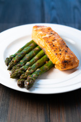 White plate with roasted salmon filet with fried asparagus on a dark wooden background