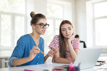 Pretty girl in eyeglasses with pencil thoughtfully working with colleague on laptop together. Young women spending time in office