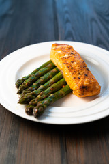 White plate with roasted salmon filet with fried asparagus on a dark wooden background