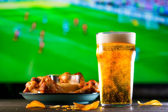 A glass of beer and hot chicken wings on a dark wooden table with crushed potato chips. Football on a background, high resolution
