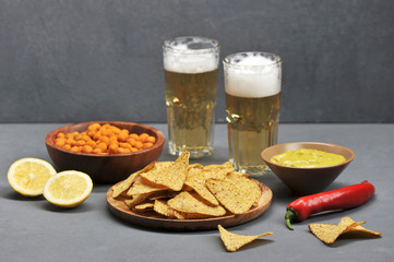 Two glasses of light beer and snacks. On the plates, peanuts in a crispy spicy crust, corn chips nachos, Mexican guacamole sauce. Next to the plates are two halves of lime. Gray background.