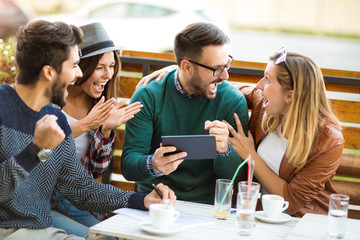 Group of four friends having a coffee together. Two women and two men at cafe talking laughing and enjoying their time using digital tablet.
