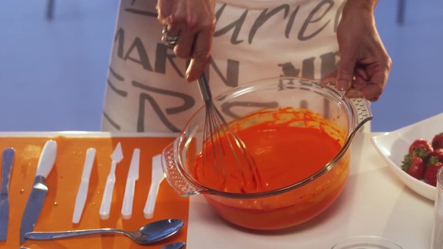 Woman's hand is whisking orange substance in big glass bowl with wide edges, put on white table with set of different sized knives and two metallic spoons plus other cooking stuff.