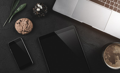 Modern laptop, mobile phone, tablet, coffee cup and biscuit on black stone background. Top view with copy space, flat lay. Light effect in the top corner.