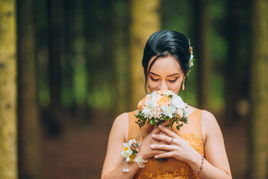 Portrait of a beautiful bridesmaid posing with bride's wedding bouquet in pine forest. Woman wear in orange long dress.