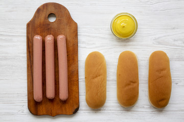 Ingredients for making hot dogs: sausages on wooden board, hot-dog buns and mustard on white wooden background, top view. Flat lay, overhead, from above.