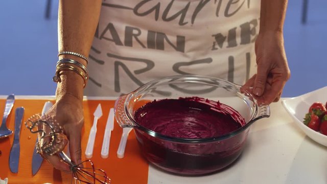 Process of hand whisking of violet substance in big round shaped glass pan with wide edges put on white table with set of defferent plastic knives, orange napkin and other cooking stuff.