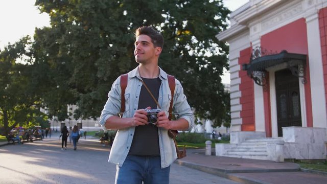 Attractive young handsome man tourist with backpack walking in city center and looking around during sunset, slow motion