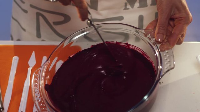 Process of woman's hand whisking of violet substance in big round shaped glass pan with wide edges put on white table with set of defferent sized knives, orange napkin and other cooking stuff.