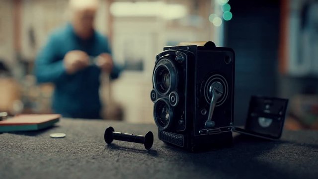 An old man comes in focus and repairs an old vintage camera on his desk, many old vintage cameras in showcase