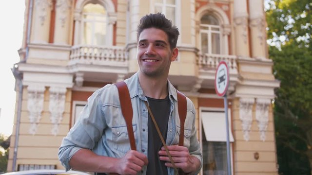 Portrait of young smiling attractive man tourist with backpack in city center, slow motion, low angle view