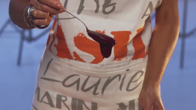 Woman wearing apron is holding spoon with violet substance dripping into glass pan with wide edges, put on white table with set of different sized knives, whisks, orange napkin and other cooking stuff