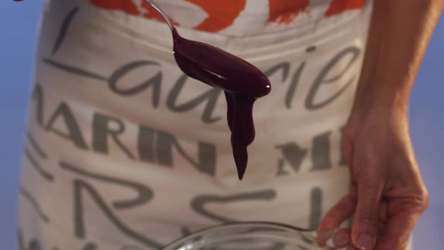 Process of violet substance smoothly spilling over metallic spoon and then dripping down with blurry colofrul bright background of woman silhouette wearing white and grey apron.