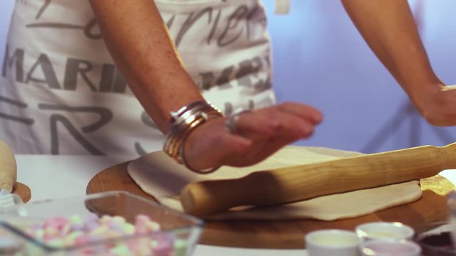 Woman wearing white and greay apron is rolling out dough on large round shaped wooden board with rolling pin on white kitchen table with defferent cooking stuff.