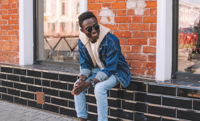 Fashion smiling african man wearing jeans jacket sits on city street, brick wall background