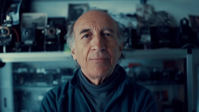 An old Caucasian man smiles at the camera in his shop of vintage cameras