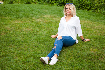 Fototapeta premium beautiful young smiling girl in sunglasses, white blouse and blue jeans sits on the grass