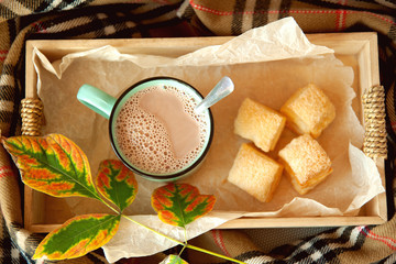 Cocoa with milk in a ceramic mug and puff pastry in a wooden box, surrounded by a checkered scarf.