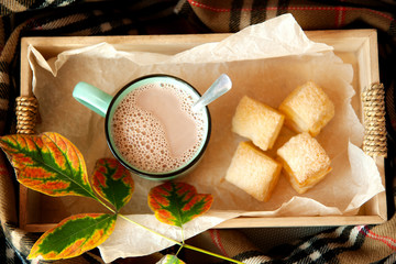 Cocoa with milk in a ceramic mug and puff pastry in a wooden box, surrounded by a checkered scarf.