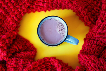 Cocoa with milk in a blue ceramic mug, on a yellow background, surrounded by a red knitted scarf