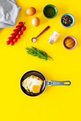 Ingredients for fried eggs with vegetables. Ready eggs in a frying pan near cherry tomatoes, greenery, spices, raw eggs on yellow background top view