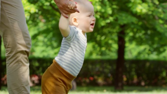 Tracking shot with tilt up of unrecognizable mother holding hands of baby boy and helping him walk on green grass outdoors