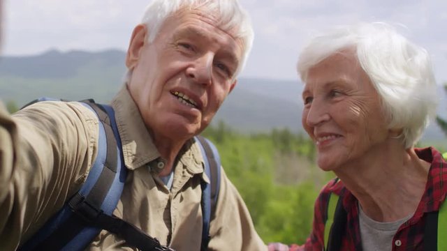 Happy senior couple smiling and telling about hike at camera while standing at viewpoint in mountains