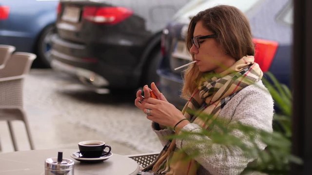 Woman lights up cigarette sit smoking at the street cafe table while drinking coffee