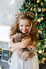 little sweet girl sits under a Christmas tree with a soft toy teddy bear