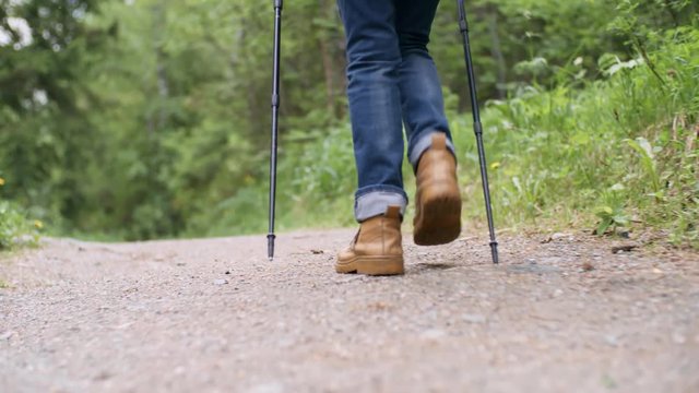 Close-up following shot of legs of female backpacker in hiking boot and jeans walking on path in forest with trekking poles