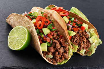 Mexican tacos with beef, tomatoes, avocado, chili and onions on a shale board background.