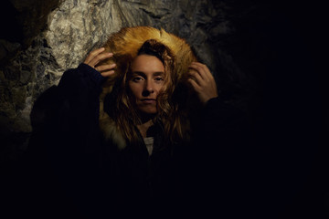 Authentic close up portrait of Caucasian female traveler, dressed in dark coat with foxy fur hood, hiding in cave from thunder, spending vacations in mountains hiking. Tourism and exploring concept.
