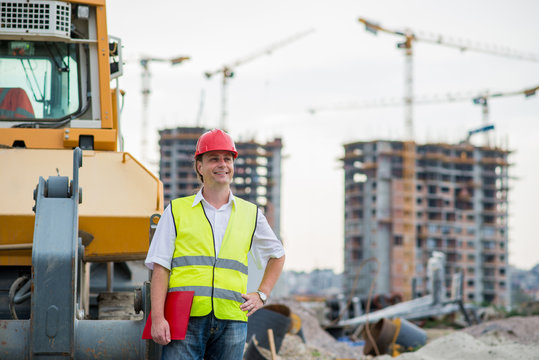 Happy engineer in front of excavator on a construction site with buildings and cranes in the background