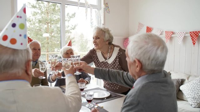 Joyous senior woman in party hat saying thanks to guests at celebration table, smiling and toasting with champagne while hosting birthday party at home