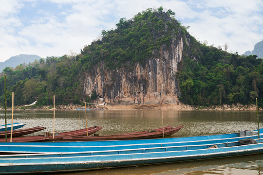 Moored boats on the Mekong River in front of a limestone cliff where the famous Pak Ou Caves are set. They are located near Luang Prabang in Laos.