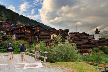 Tourists walk through the beautiful alpine village Grimentz, Switzerland, with traditional wooden houses and church tower in summer 