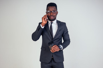 Handsome young African man in formal black suit holds a plastic paper cup with hot coffee cocoa (tea) to go and smartphone. businessman on coffee break after meeting on white background in studio