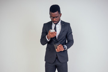 Obraz na płótnie Canvas Handsome young African man in formal black suit holds a plastic paper cup with hot coffee cocoa (tea) to go and smartphone. businessman on coffee break after meeting on white background in studio
