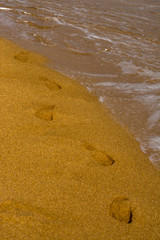 A string of bare footprints in the sand. Waves on the sea coast, sandy beach. Foam on sea water.