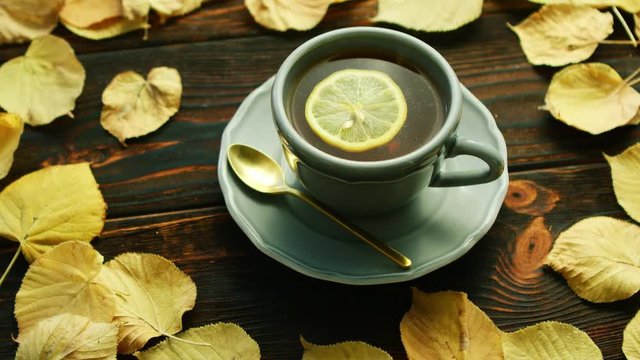 From above view of cup of hot tea with slice of lemon with saucer and spoon placed on wooden background and decorated with dry yellow leaves.