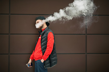 Vape teenager. Young handsome bearded white man in black cap smoking an electronic cigarette and letting off the steam opposite the modern brown background in autumn.