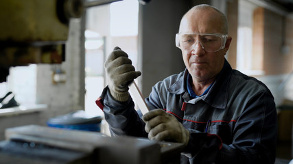 aged male worker is adjusting machine in a workshop of factory, rotating key and using hammer