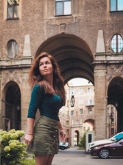 Fototapeta premium Brunette girl in a skirt and with long hair, standing, posing in the courtyard of a historic building in St. Petersburg, the entrance of the house, the house is made of numerous huge arches