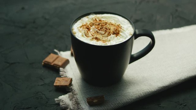 From above view of black mug of coffee with whipped cream on top and pieces of chocolate laid near on gray background
